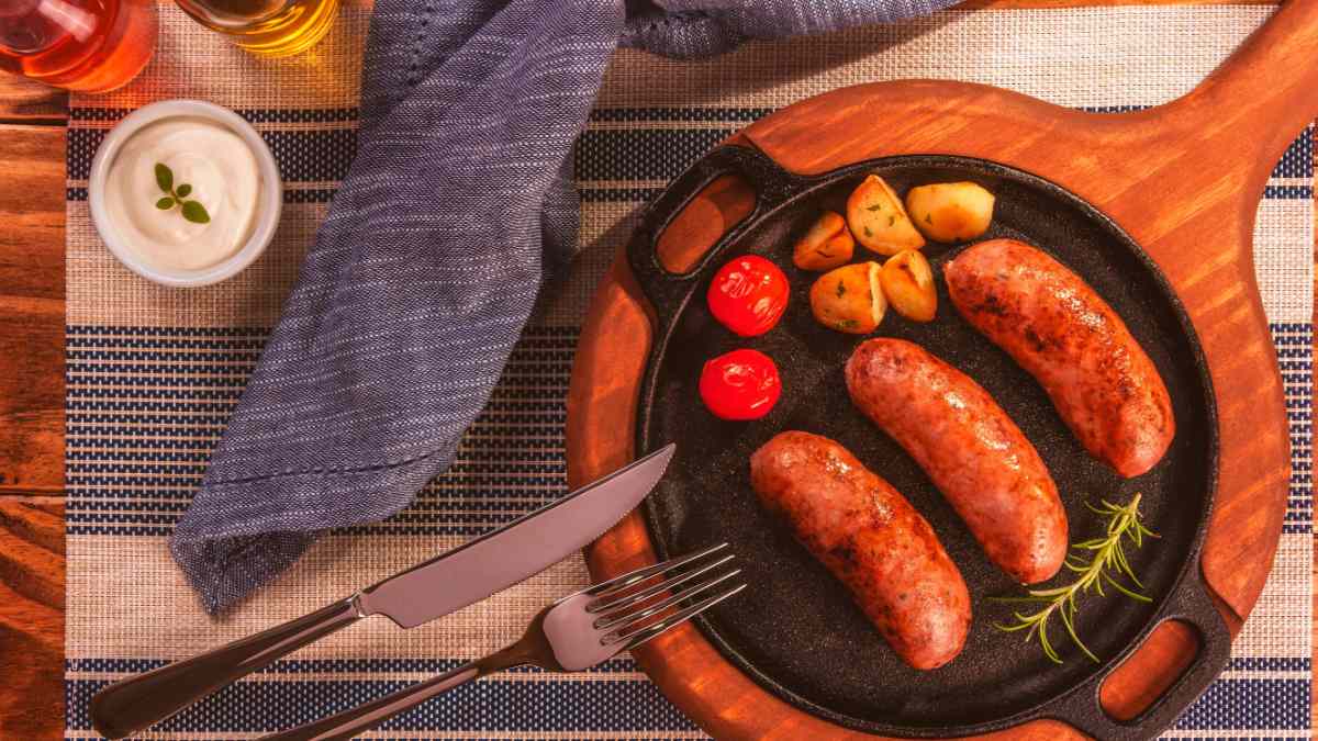 Mesa posta com uma toalha azul e branca. Ao lado direito uima tábua de amdeira com uma pedra ao centro com 3 linguiças, tomates e batatas. AO lado esquerda talhares e um potinho com molho.