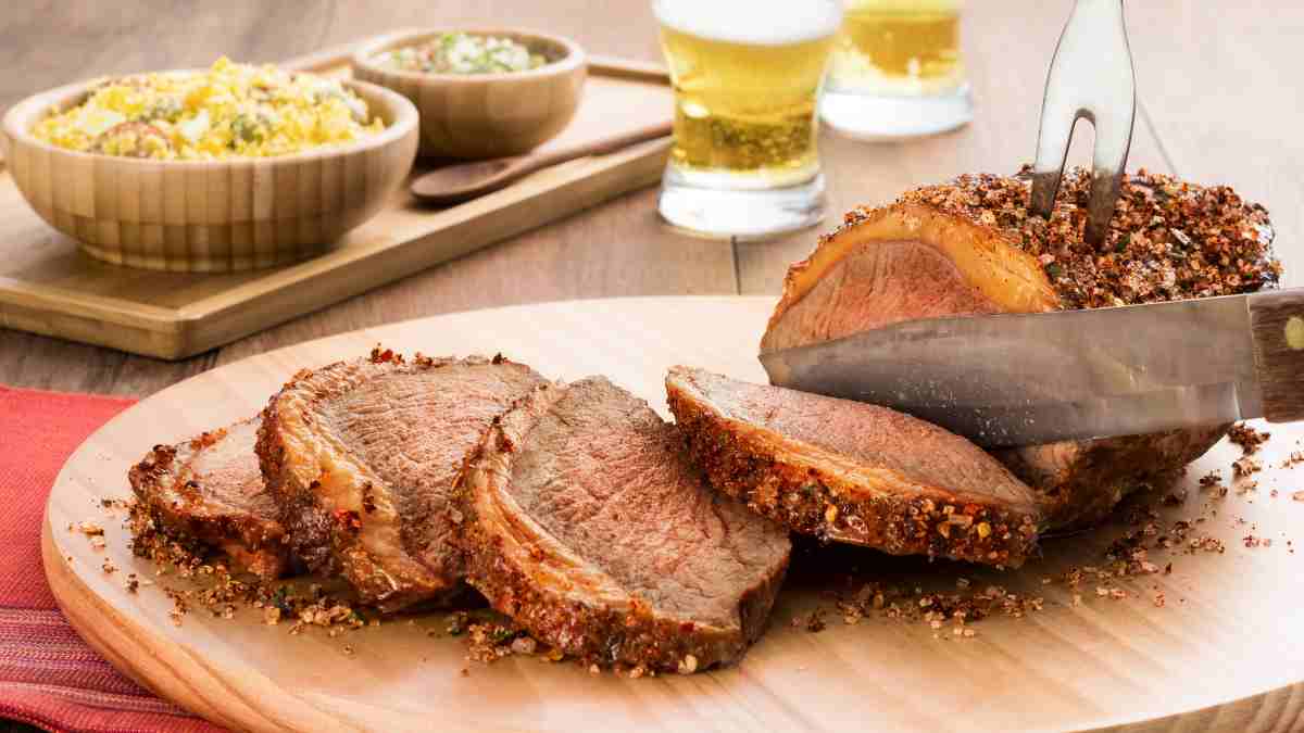 Churrasco de picanha sendo fatiada em uma tábua de madeira redonda clara. Atrás da tábua, alguns potes de molhos para acompanhar.