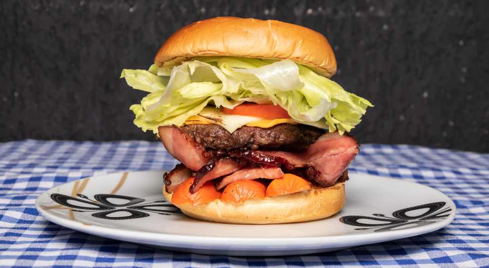 X Tudo brasileito srvido com salada de alface, hambúrguer bovino, ovo frito, fatias de bacon e de salsicha e rodelas de tomate no pão brioche, em um prato branco redondo e raso com dsenhos pretos, em cima de uma mesa com um pano quadriculado branco e azul