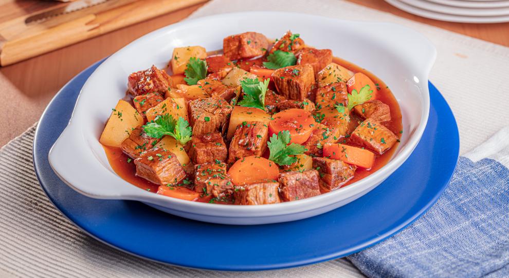 A imagem destaca uma travessa redonda de porcelana com guisado preparado com cubos de carne, legumes em cubos e folhas de coentro. A travessa redonda está sobre um prato redondo azul e raso.