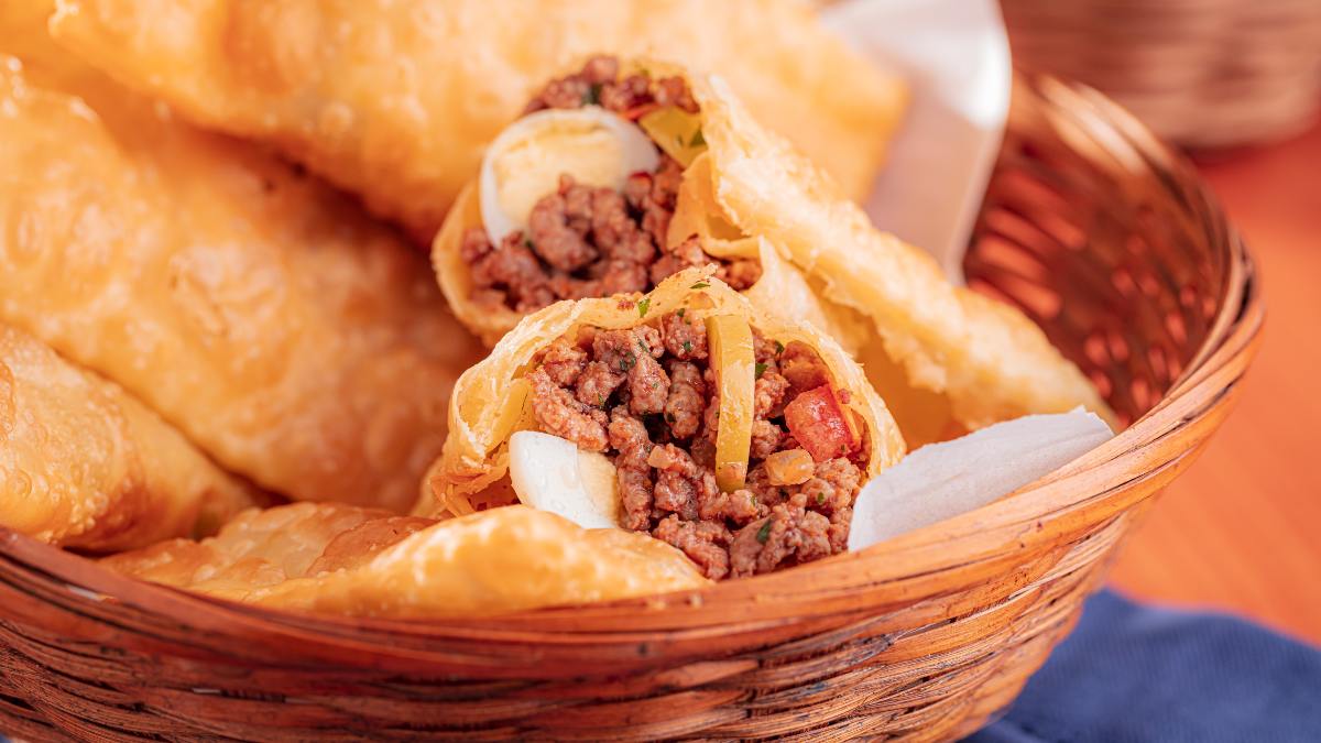 A imagem destaca em cesta com pasteis de carne moída. A imagem estpa com foco em um dos pasteis que está partido no meio, com recheio de carne moída, ovo de codorna e tomates picados.