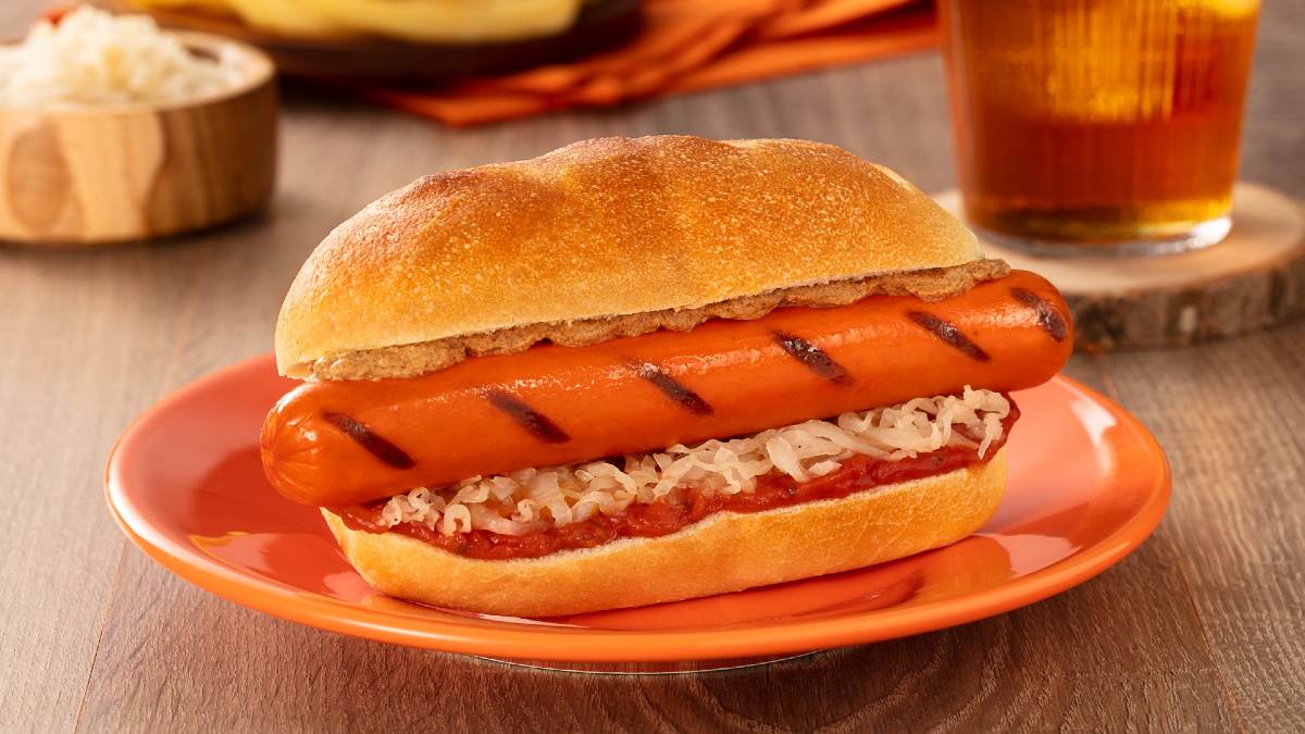 A imgem destaca um cachorro quente, servido em um prato laranja no centro de uma mesa de madeira.