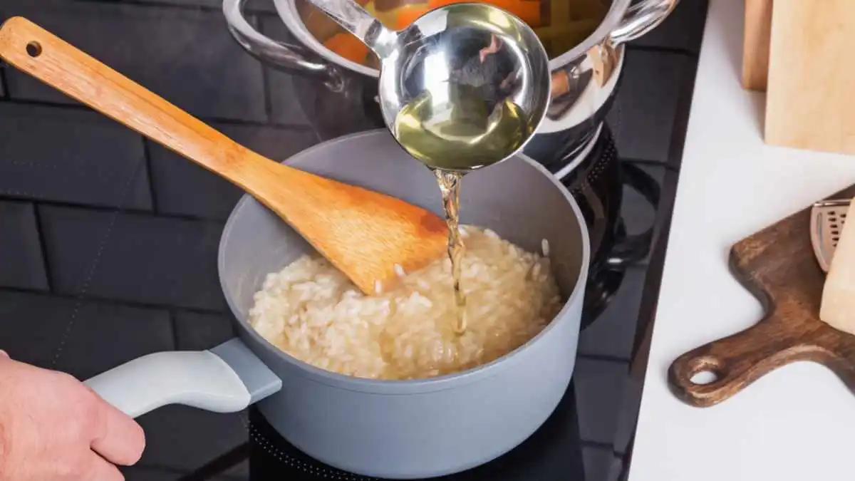 Imagem destaca a mão de pessoa branca segurando uma panela, ao preparar um risoto de carne. Na imagem, a pessoa está adicionando caldo de legumes aos poucos para incorporar o risoto.