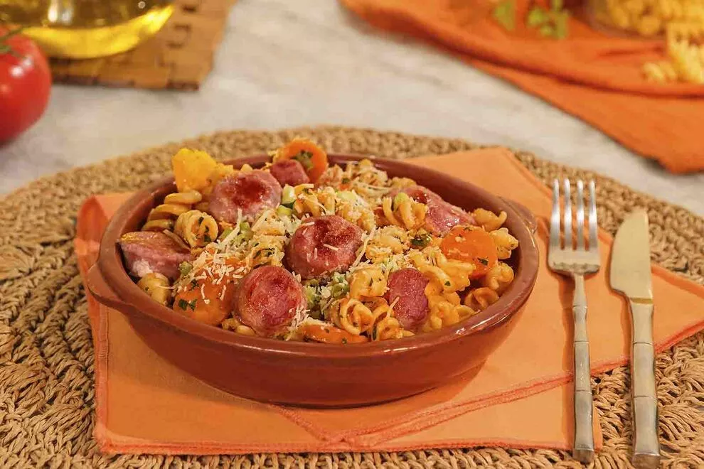 Imagem de um prato fundo de cerâmica com Macarrão de Panela de Pressão com Linguiça, em cima de uma toalha branca laranja, sob uma jogo de mesa de corda trançada, em cima de uma mesa de mármore, com um par de talheres ao lado