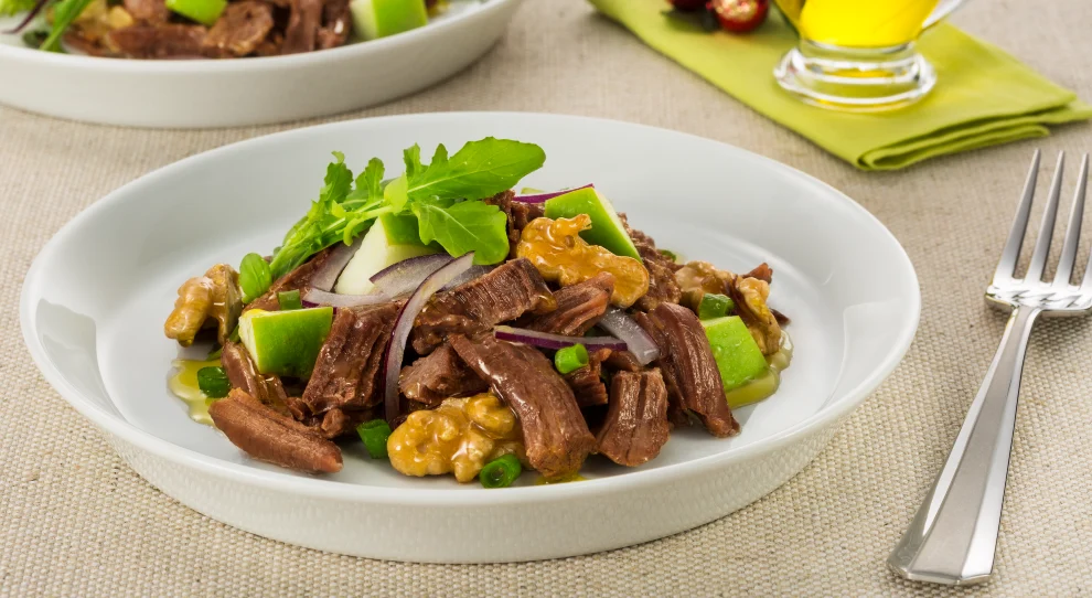 A imagem mostra dois pratos brancos, cada um contendo uma salada de carne em tiras. A salada é composta por pedaços de carne suculenta, folhas de rúcula, nozes, fatias de maçã verde, tiras de cebola roxa e pequenos pedaços de pimentão verde. Ao lado do prato principal, há um garfo de prata. No fundo da imagem, há um segundo prato de salada semelhante e um pequeno recipiente de vidro com azeite de oliva, colocado sobre um guardanapo verde-claro. O cenário é uma mesa coberta por uma toalha de tecido bege, criando um ambiente acolhedor e elegante para a refeição