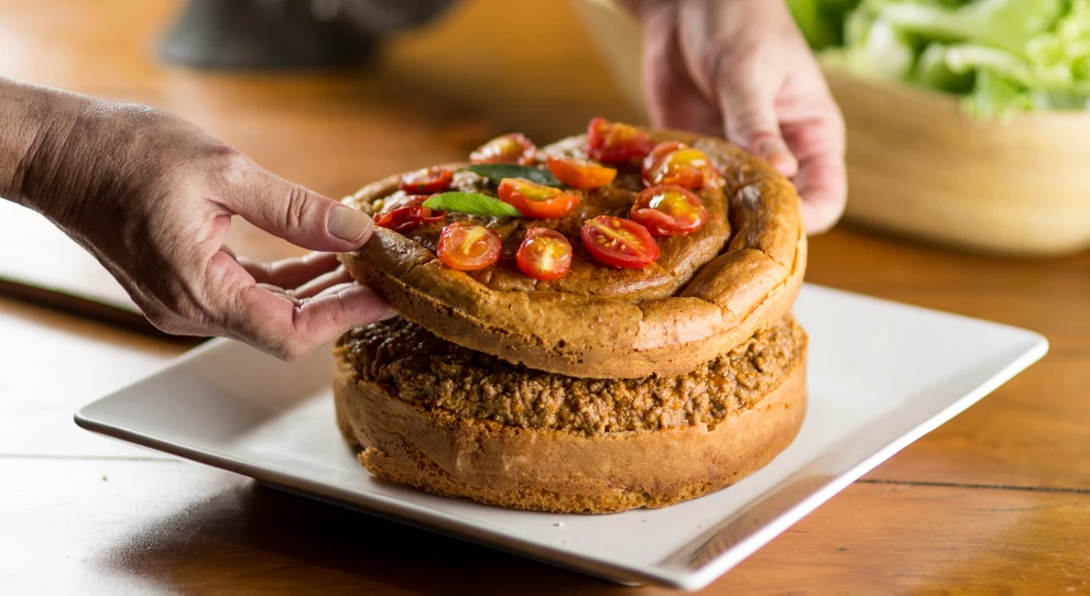 Na imagem, há uma pessoa montando uma torta mexicana de carne. A torta está sendo colocada em um prato quadrado branco sobre uma mesa de madeira. A pessoa está posicionando a camada superior da torta, que parece ser uma massa espessa e dourada, coberta com rodelas de tomate cereja frescos e folhas de manjericão.

A camada inferior da torta, já no prato, é composta por uma massa semelhante, com um recheio abundante de carne moída temperada, visível na parte superior da camada. Ao fundo, desfocado, há uma tigela de madeira contendo folhas verdes de alface. A cena tem um toque caseiro e acolhedor, com foco na preparação cuidadosa do prato.