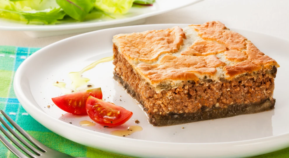 Na imagem, há uma fatia de torta grega de acém em um prato branco. A torta tem uma crosta dourada e crocante no topo, com um recheio espesso e suculento de carne moída temperada. A base da torta parece ser feita de uma camada fina e mais escura. Ao lado da fatia de torta, há alguns pedaços de tomate cereja cortados ao meio, regados com um fio de azeite e temperados com sal e pimenta. No fundo, parcialmente desfocado, vê-se um prato com folhas verdes de alface e outras verduras. O cenário é complementado por um guardanapo colorido em tons de azul e verde, sobre o qual está apoiado um garfo prateado.