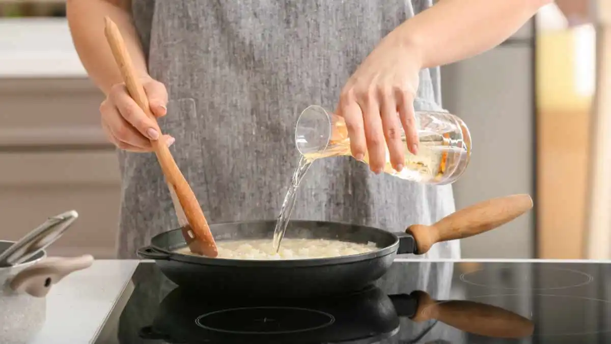 Imagem destaca uma mulher branca, de avental, adicionando vinho branco na receita de risoto de carne.