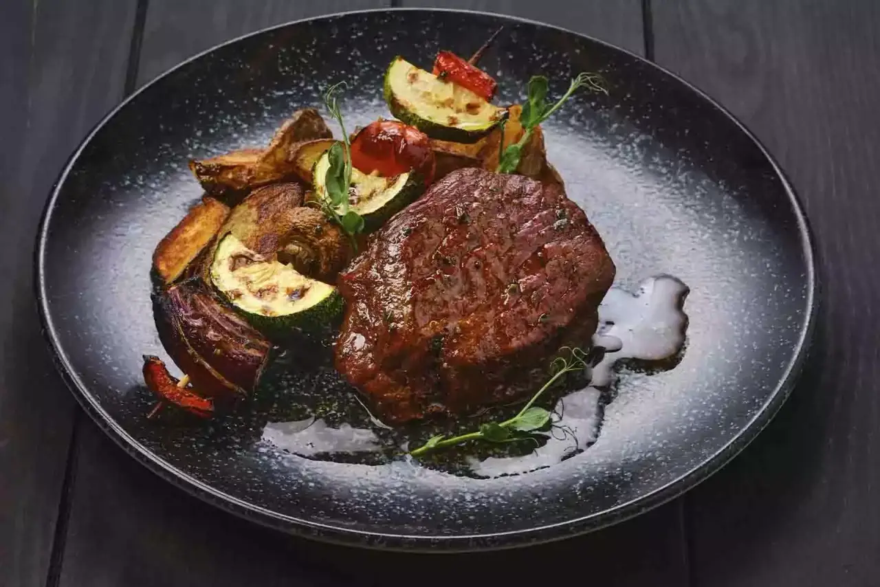 Imagem de um bife ancho assado com diversos legumes assados ao lado, sob um prato escuro redondo, em cima de uma mesa de madeira escura 