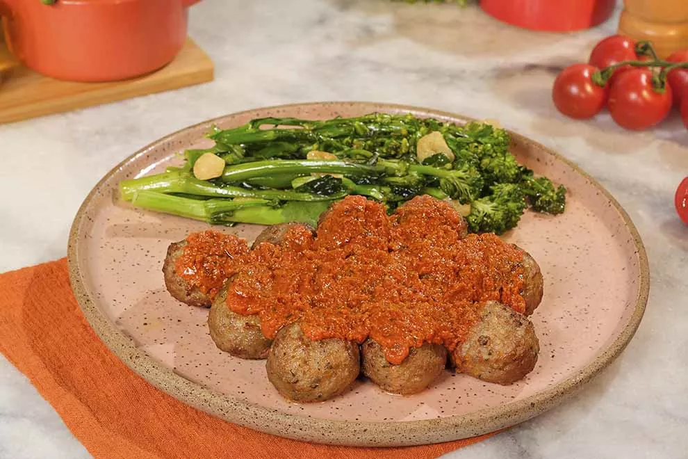 Imagem de Almôndegas assadas com salada de brócolis sob um prato redondo de cerâmica, em cima de uma mesa de mármore  