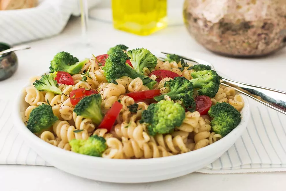 Imagem de um macarrão com brócolis e tomates cortados, em cima de um prato redondo branco, sob uma toalha listrada branca, em cima de uma mesa branca com um garfo ao lado 