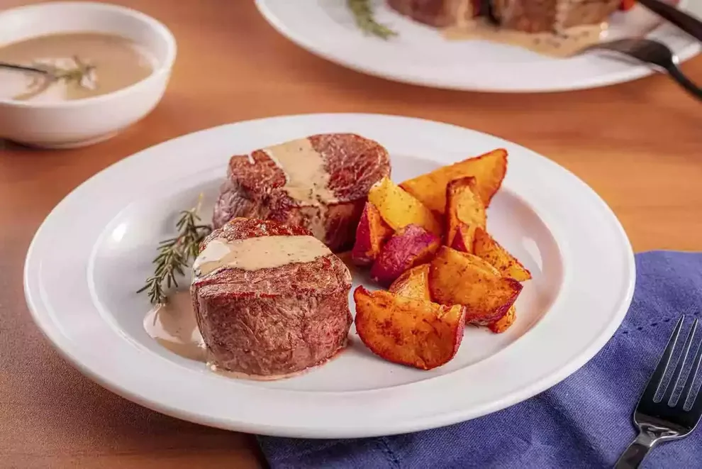 Imagem de duas peças de medalhão de mignon assadas, com um molho de gorgonzola por cima e ao lado batata doce assadas, sob um prato redondo branco, tendo um garfo ao seu lado e ao fundo uma combuca de molho de gorgonzola