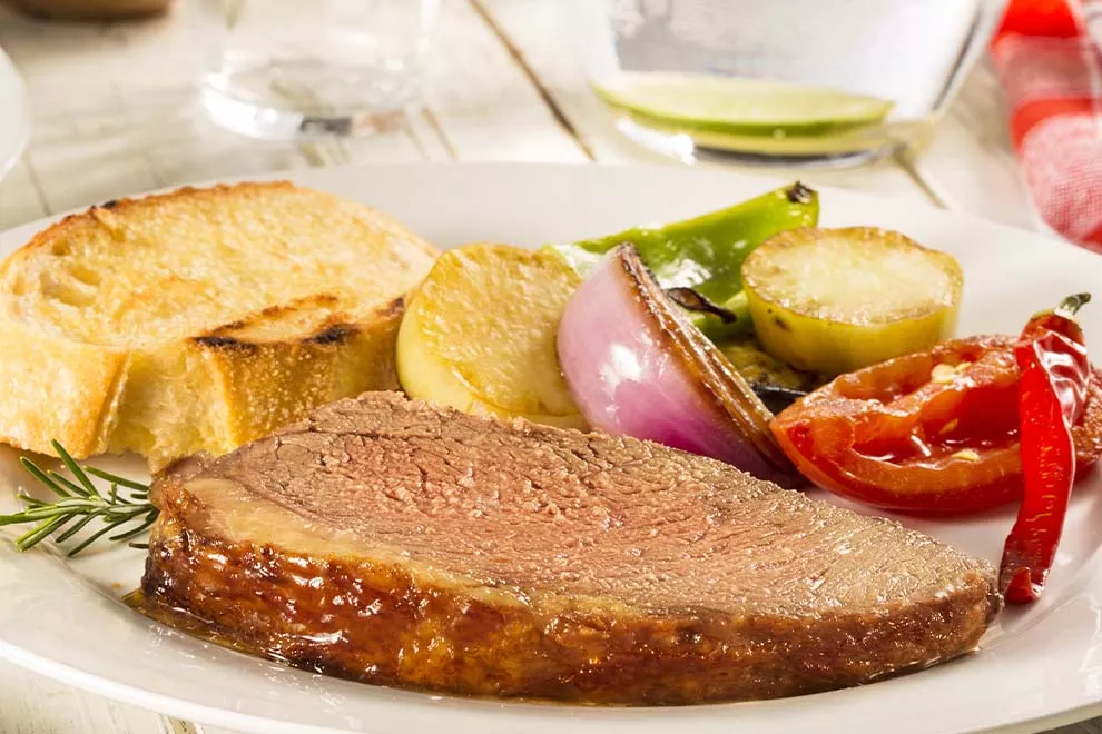 Imagem de uma fatia de picanha feita na chapa, com uma fatia de pão e legumes assados ao lado, sob um prato redondo branco, em cima de uma mesa de madeira branca 