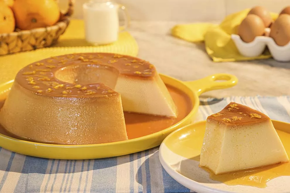 Imagem de uma fatia de pudim de pão em cima de um prato redondo amarelo, com uma travessa redonda amarela com o pudim de pão ao fundo, sob um pano listrado azul, em cima de uma mesa de mármore 