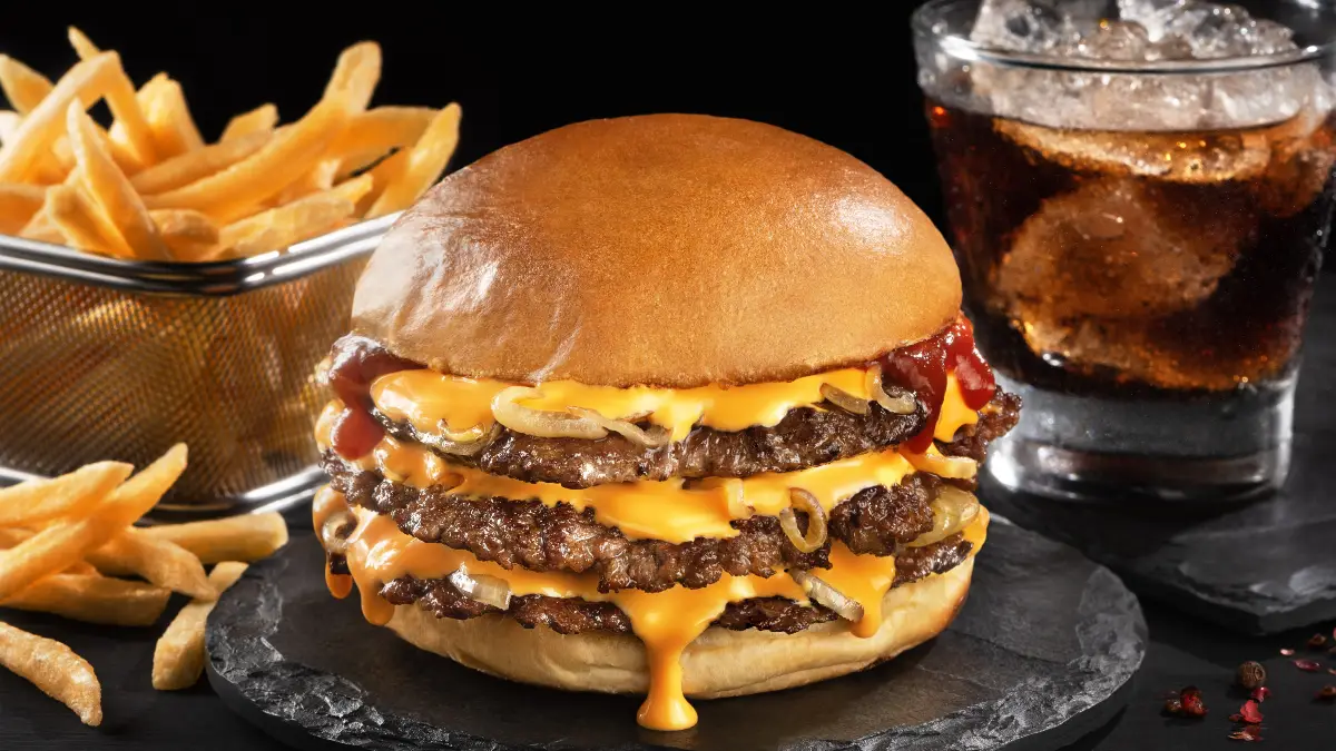 A imagem mostra um "smash burger" triplo com uma aparência irresistível. O sanduíche é composto por três hambúrgueres de carne finos e grelhados, todos cobertos com queijo cheddar derretido, que escorre pelas bordas de cada hambúrguer. Entre as camadas de carne e queijo, há anéis de cebola caramelizada e ketchup. O hambúrguer é montado em um pão macio e dourado. À esquerda do sanduíche, há uma porção de batatas fritas douradas servidas em um recipiente de metal. À direita, há um copo de refrigerante com gelo, completando o conjunto clássico de um "smash burger" com batatas fritas e bebida. A imagem é capturada em uma superfície de pedra escura, que contrasta com as cores vibrantes dos alimentos, destacando ainda mais a refeição.