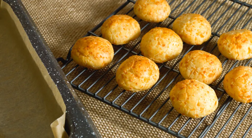 A imagem mostra uma grelha de resfriamento com pães de queijo dourados e assados, dispostos lado a lado. Os pães de queijo possuem uma casca dourada e levemente craquelada, com uma textura que aparenta estar crocante por fora e macia por dentro. Ao lado esquerdo da grelha, há uma forma forrada com papel manteiga, indicando que os pães acabaram de sair do forno. A superfície embaixo da grelha é feita de tecido rústico de cor bege. A iluminação natural destaca o brilho dos pães, dando uma sensação de frescor e sabor.